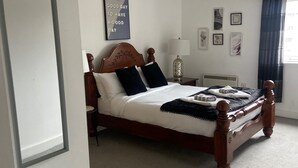 Un lit bien fait, avec une tête de lit en bois, une table de chevet avec une lampe, et un mur orné de tableaux et d’une citation inspirante.
