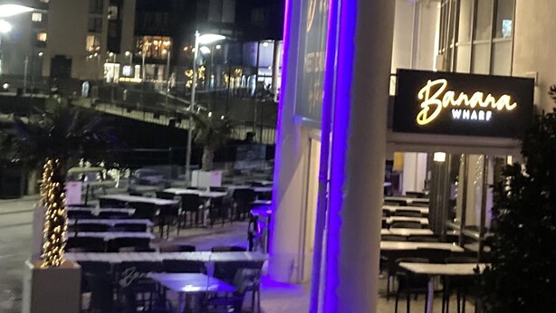 Un espace extérieur aménagé avec des tables et des chaises, une enseigne indiquant « Banana Wharf », et une vue sur un bâtiment en arrière-plan.