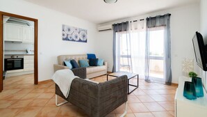 A living room with a sofa, coffee table, and a TV. There is a kitchen area with cabinets and an oven. The room has a large window with curtains. - Odissi Apartment, Vilamoura, Algarve (Quarteira)