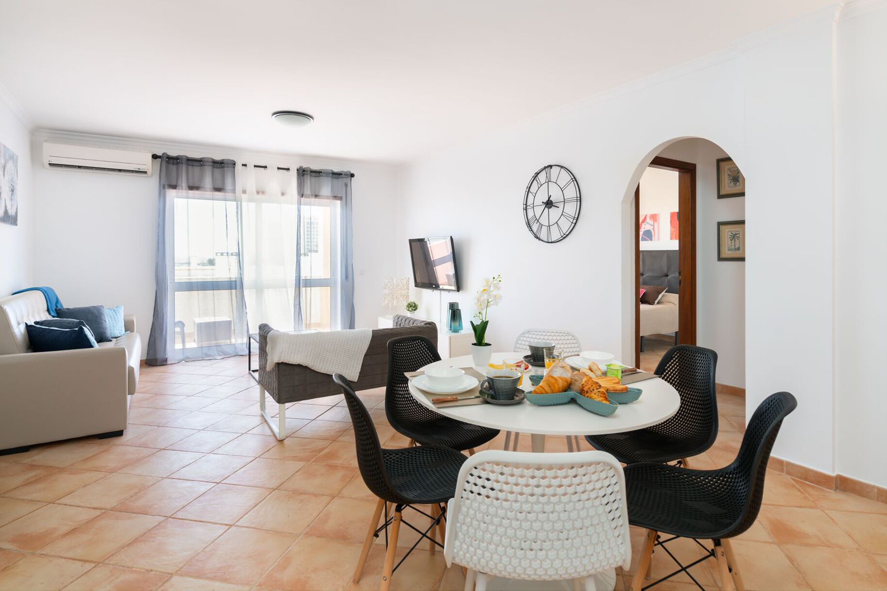 A modern living room with a dining area, featuring a round table, black and white chairs, and a large window with curtains.