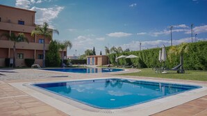 A swimming pool area with two pools, a building with multiple floors, and a well-maintained lawn with palm trees. - Odissi Apartment, Vilamoura, Algarve (Quarteira)