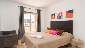 A bedroom with a bed, a chair, a window with curtains, and artwork on the wall. - Odissi Apartment, Vilamoura, Algarve (Quarteira)
