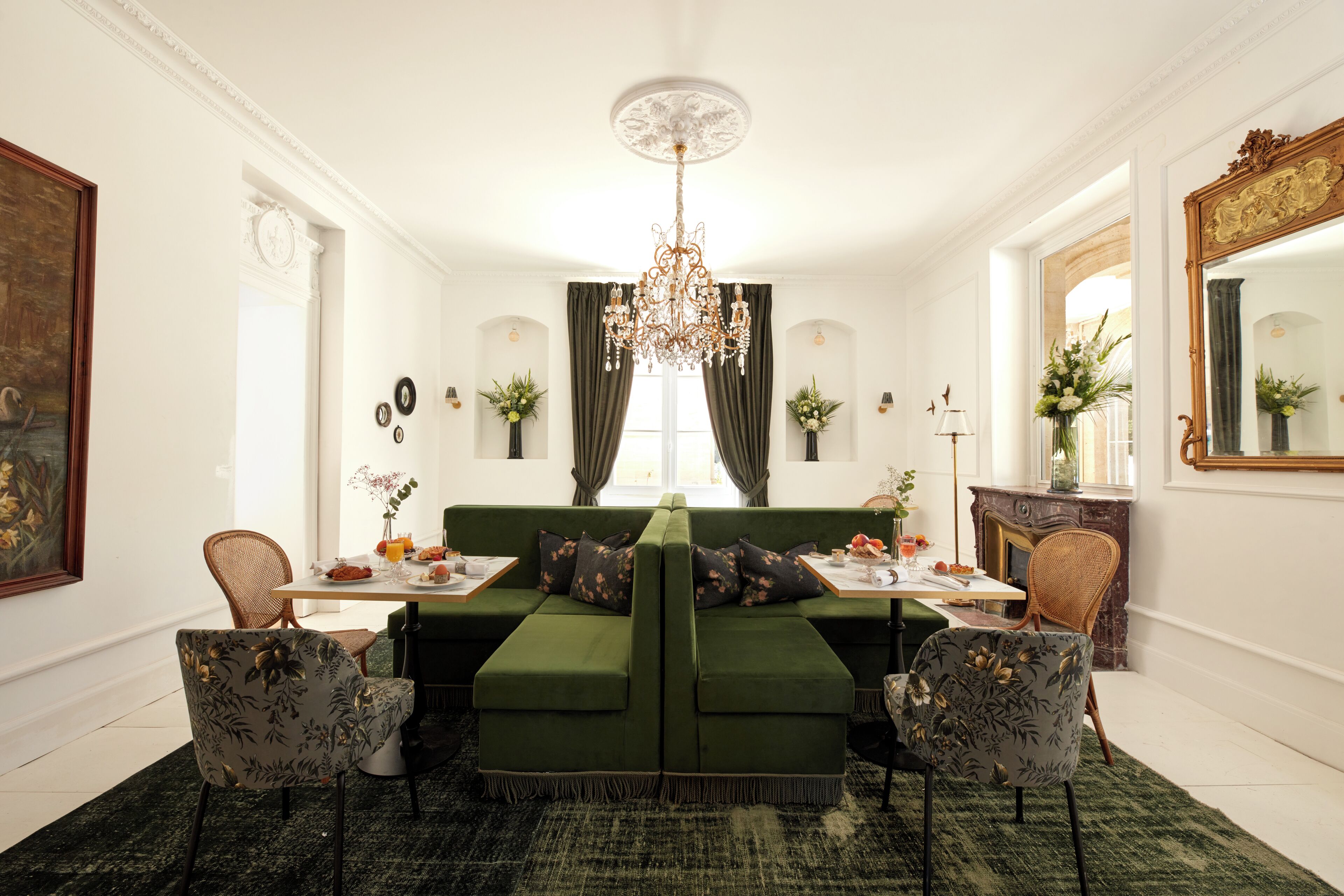 A dining area with a green sofa, two chairs, a chandelier, and a table set with plates and flowers.
