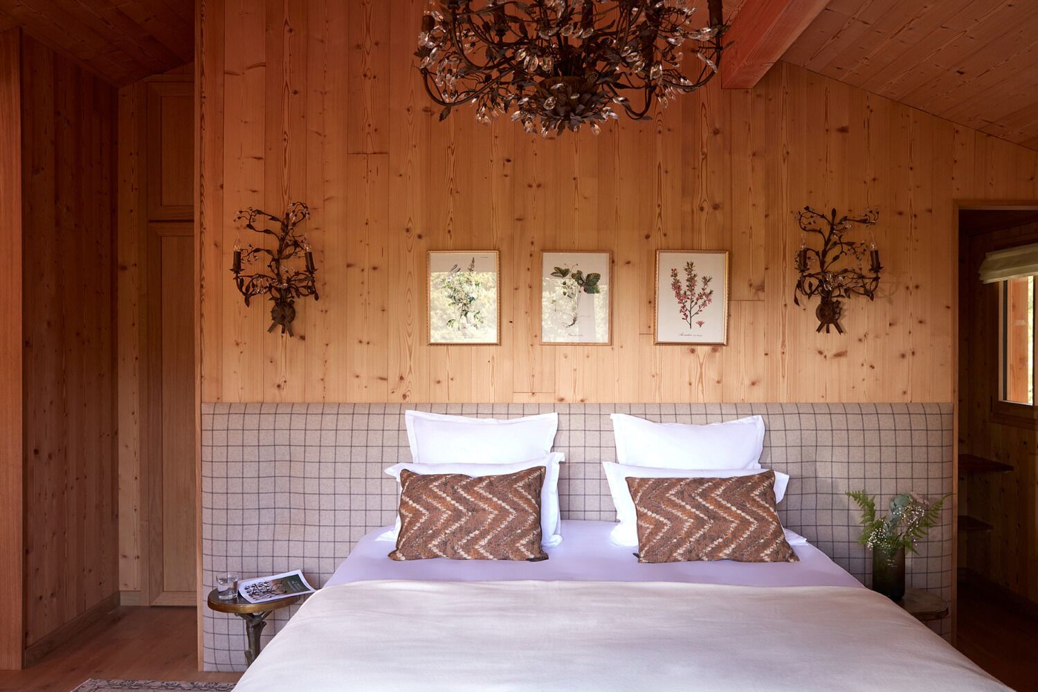 Una habitación con paneles de madera, una cama, dos impresiones enmarcadas de plantas y un candelabro.