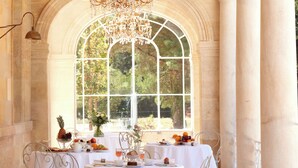 A dining area with a table set for a meal, a chandelier, and a large arched window with a view of greenery. - Château Léognan, Domaine viticole - proche Bordeaux (Leognan)