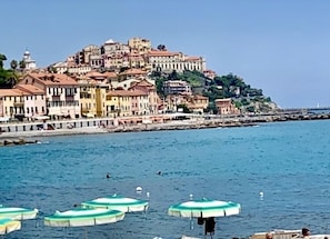 A coastal town with buildings on a hill overlooking the sea, boats with umbrellas, and a lighthouse in the distance.