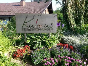 Ein Schild für 'Kuhlaus Ferienwohnungen' in einem Garten mit verschiedenen Blumen und Pflanzen.