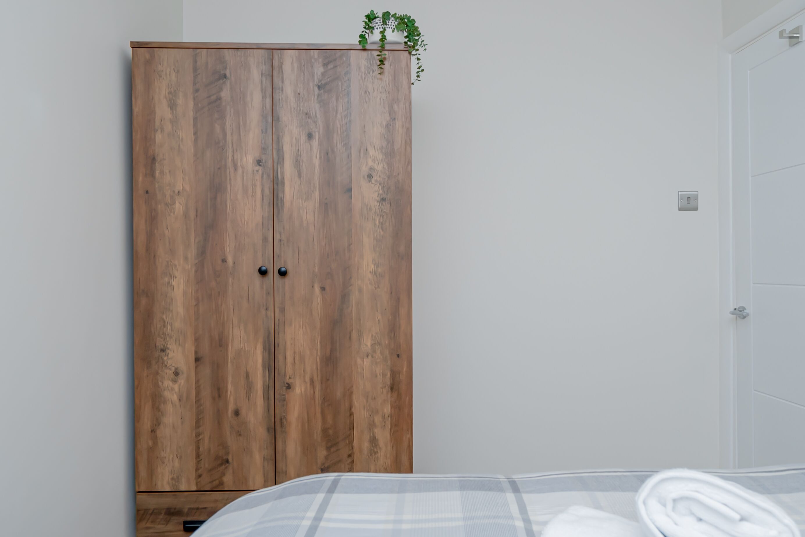 A bedroom with a wooden wardrobe, a bed with a white and grey striped bedspread, and a small potted plant on top of the wardrobe.