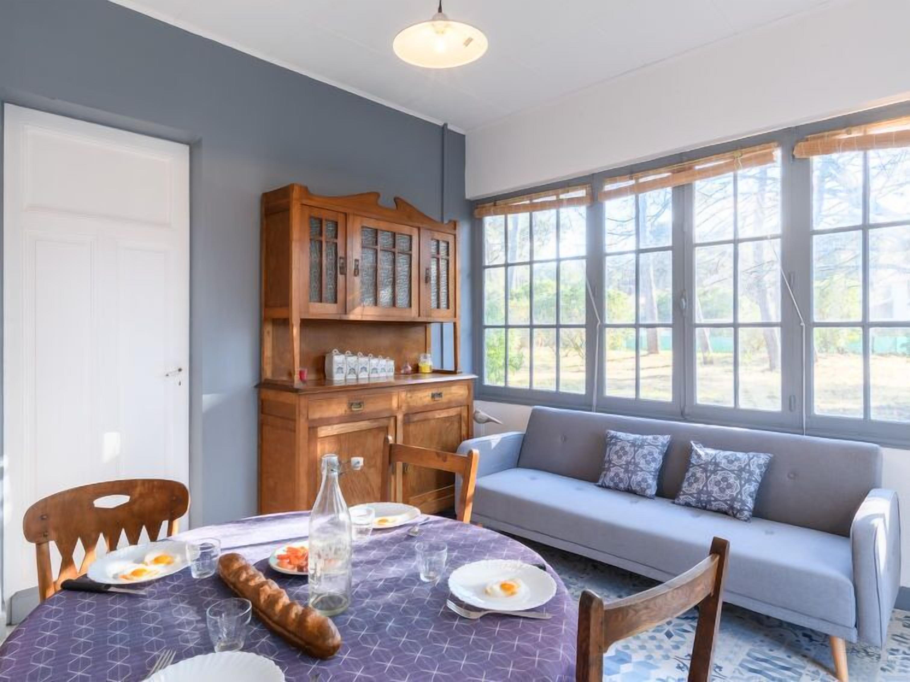 A dining area with a table set for two, a wooden cabinet with glass doors, and a sofa by a large window.