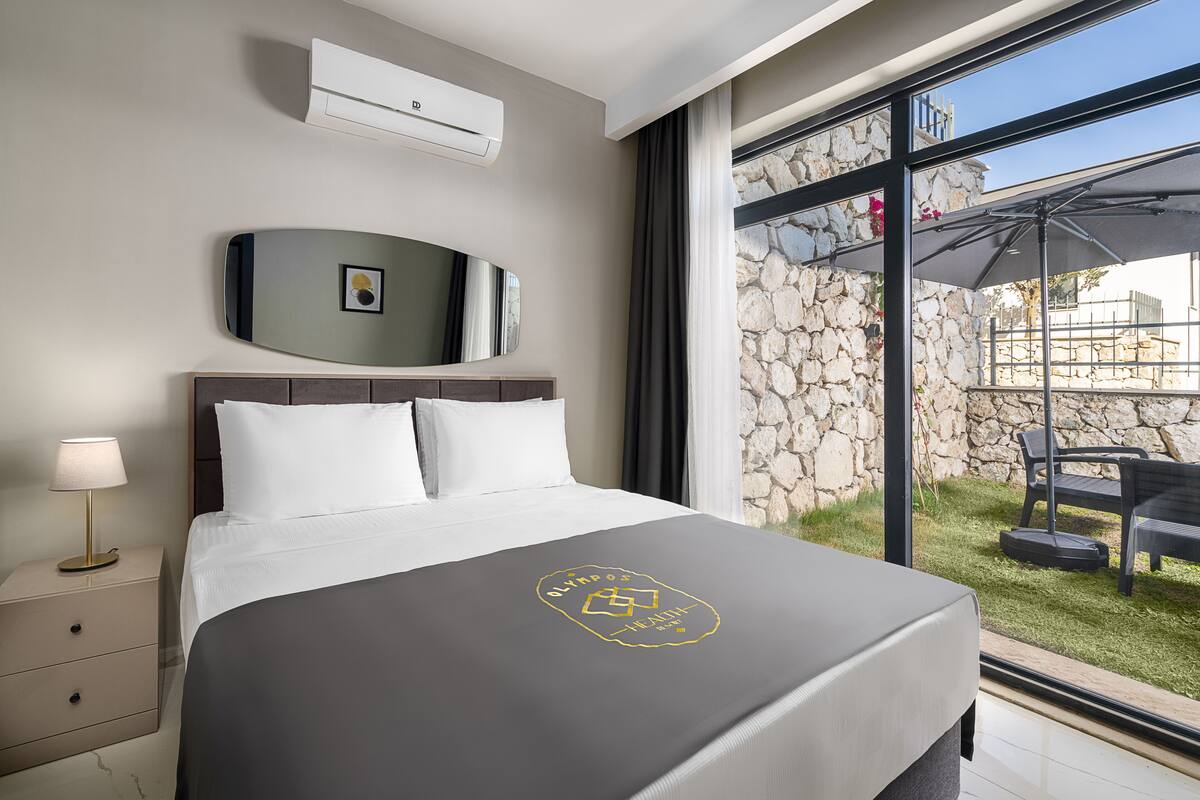 Ein Hotelzimmer mit Bett, Nachttisch, Lampe, Wandklimaanlage, einem gerahmten Bild und Blick auf eine Außenpatio mit Steinmauer und Tisch.