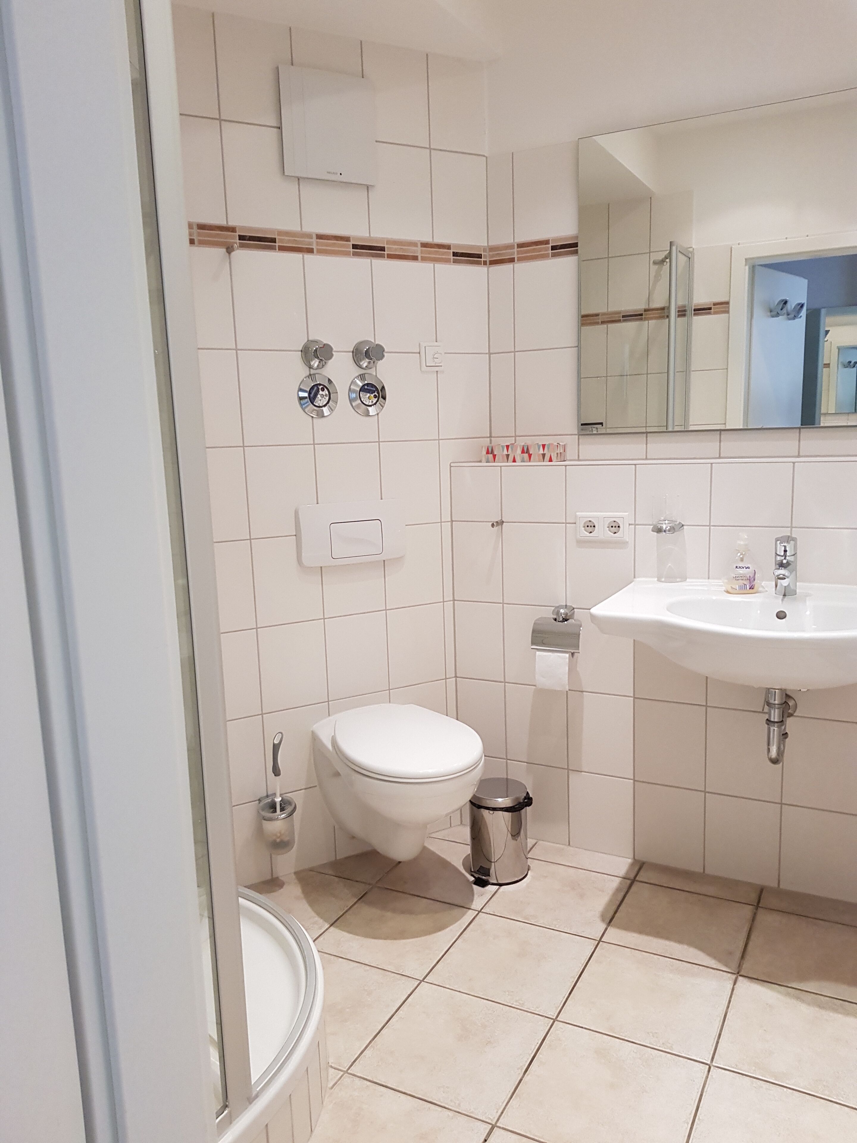 Une salle de bain avec des toilettes, un lavabo et un miroir. Il y a deux robinets fixés au mur et une poubelle.