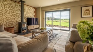A living room with a stone wall, a fireplace, a television, and a large window offering a view of a green landscape. - Hayloft, Burnham Market, Norfolk (Burnham Market)