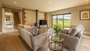 A modern living room with a stone wall, a fireplace, a large window, and a sofa. - Hayloft, Burnham Market, Norfolk (Burnham Market)