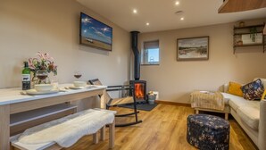 A cozy living room with a wooden floor, a sofa, a dining table with chairs, a fireplace, and a TV mounted on the wall. - Bumblebee Cottage, Culford Heath, Suffolk (Bury St Edmunds)