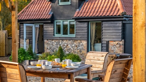 An outdoor wooden table set with breakfast items, a stone wall, and a house with a red-tiled roof in the background. - Bumblebee Cottage, Culford Heath, Suffolk (Bury St Edmunds)