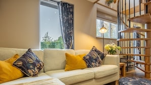 A cozy living room with a sofa, patterned cushions, a wooden staircase, and a view of greenery outside. - Bumblebee Cottage, Culford Heath, Suffolk (Bury St Edmunds)