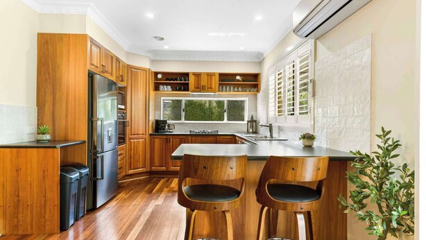 Cocina con armarios de madera, un refrigerador de acero inoxidable, horno empotrado, un lavamanos y dos taburetes de bar.