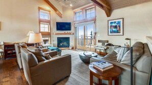 A spacious living room with a fireplace, leather sofas, a coffee table, and a large window with a view. - The Lodges D-1 - 1 Block to the Beach! (Cannon Beach)