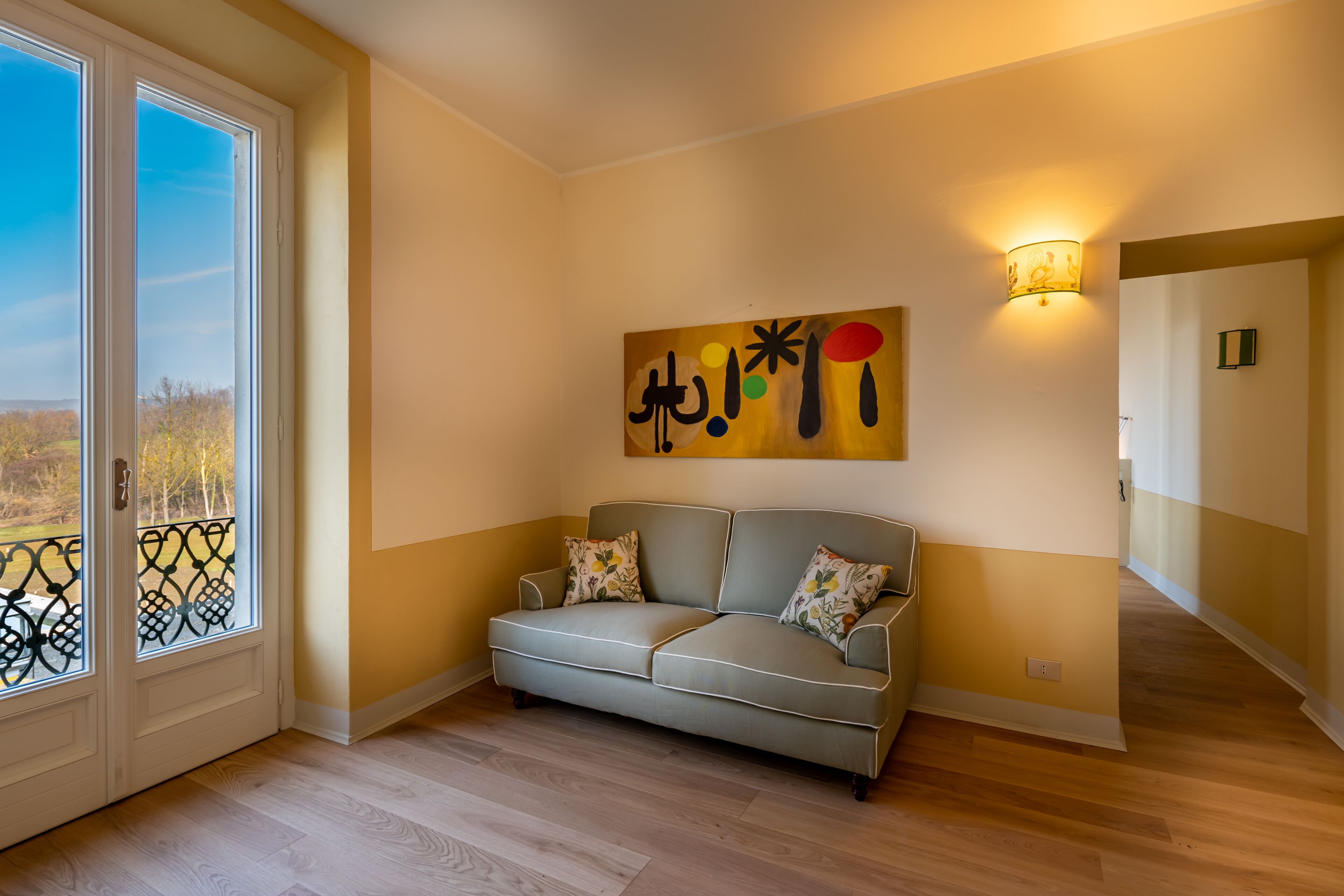 Ein Zimmer mit einem beige gefärbten Sofa, einem großen abstrakten Gemälde, einer Wandleuchte und Blick durch eine Glastür.