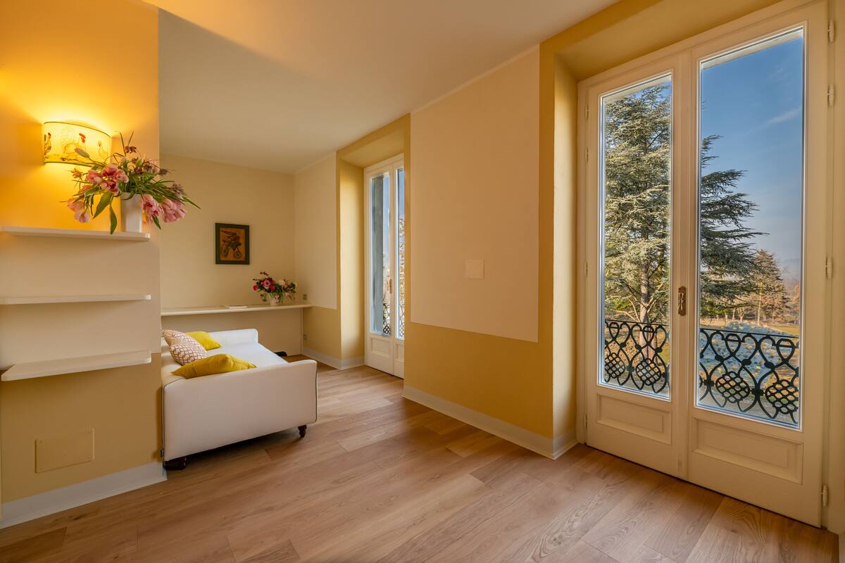 Une pièce avec un canapé blanc, une petite table sur laquelle est posé un vase de fleurs, une lampe fixée au mur et une grande fenêtre donnant sur des arbres.