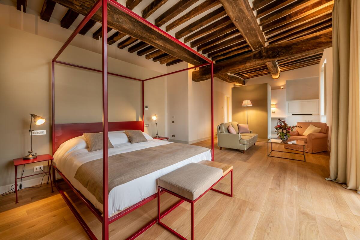 Une chambre spacieuse avec un lit à baldaquin, une table de chevet, un banc et un coin salon comprenant un canapé et un fauteuil.