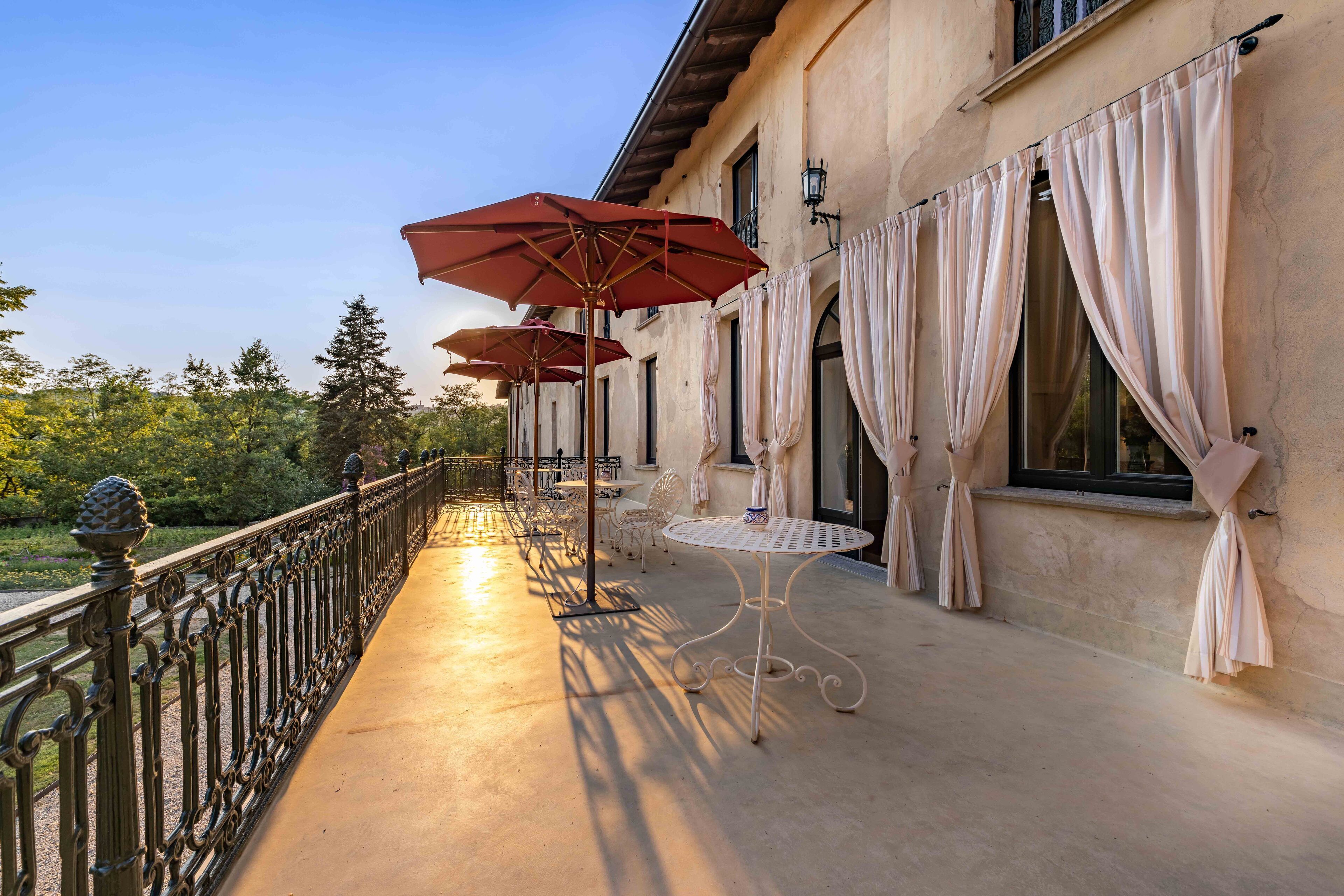 Un balcón con mobiliario exterior, incluyendo mesas y sombrillas rojas, con vistas a un jardín.