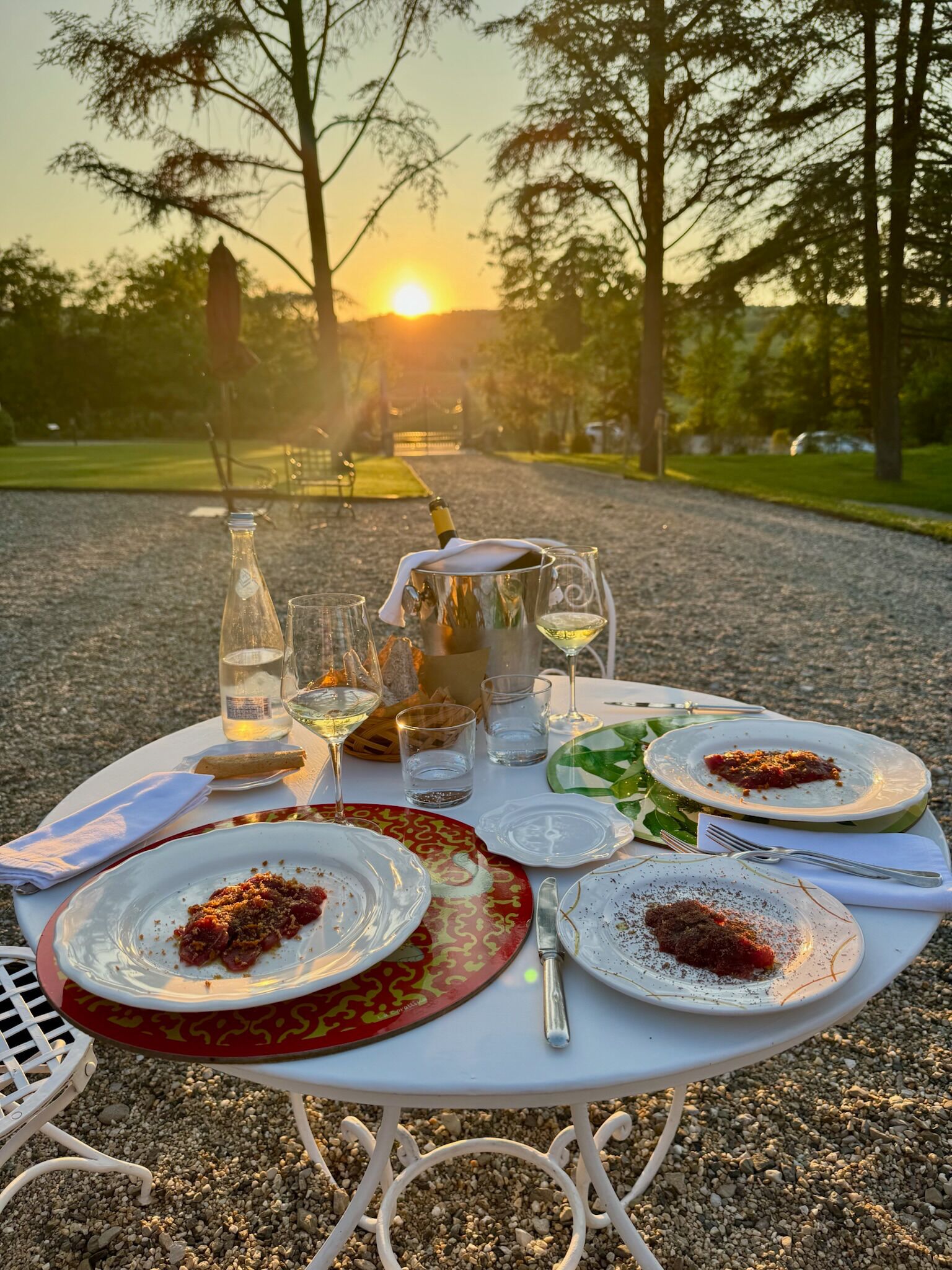 Ein Esstisch im Freien für zwei Personen, gedeckt mit Speisen, Gläsern und einer Weinflasche, mit Sonnenuntergang im Hintergrund.