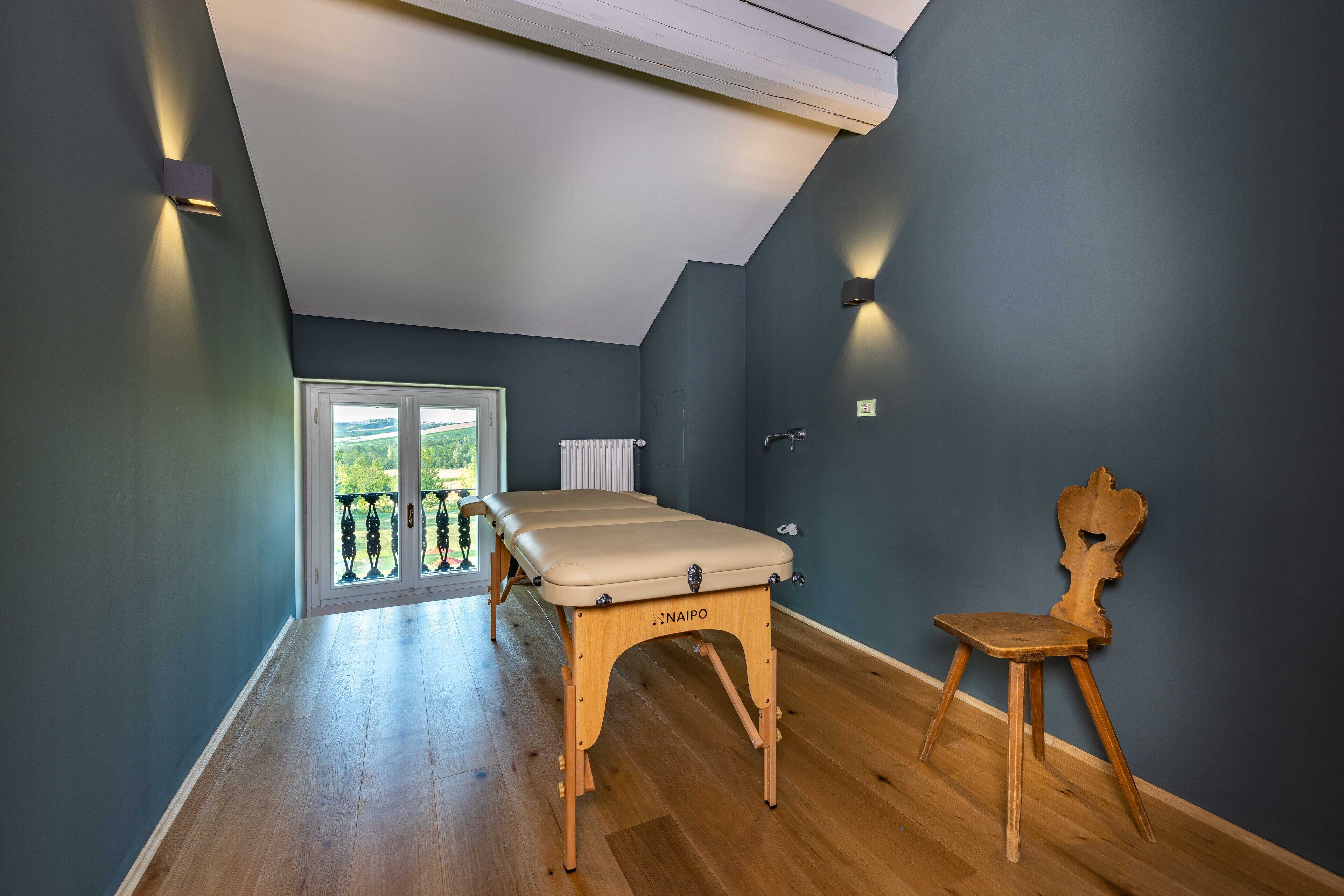 A room with a wooden floor, a massage table, a wooden chair, and a view through a glass door.
