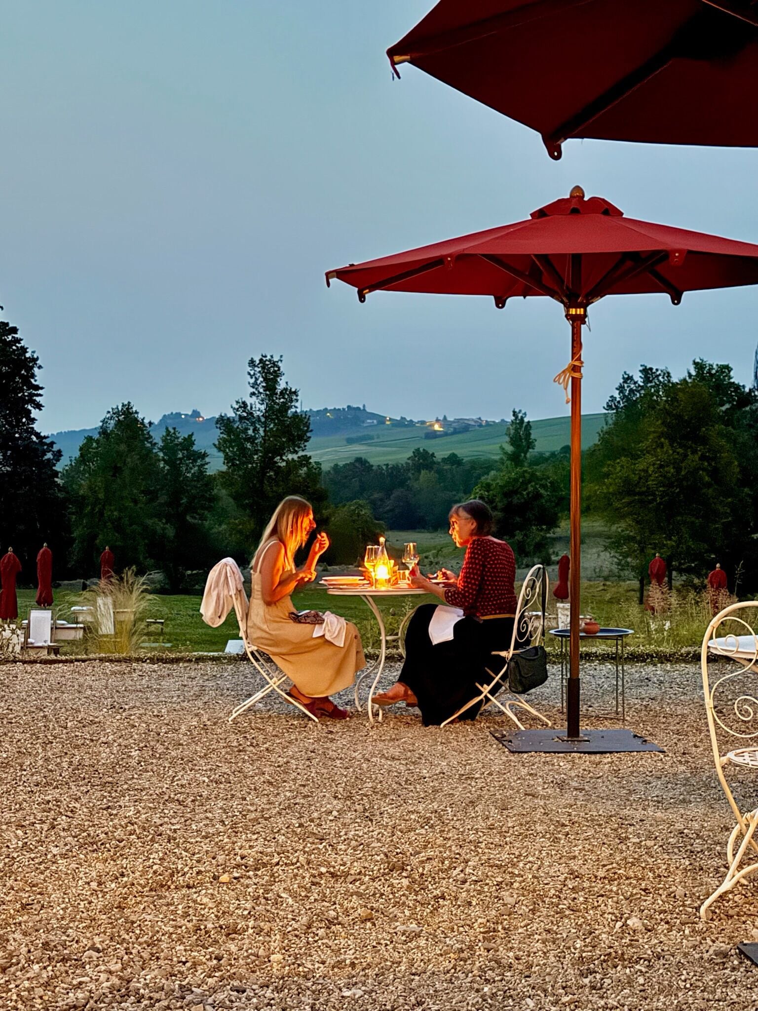 Zwei Personen speisen an einem Tisch im Freien, der mit einem roten Sonnenschirm ausgestattet ist, umgeben von Kies und Pflanzen, mit Blick auf Hügel im Hintergrund.