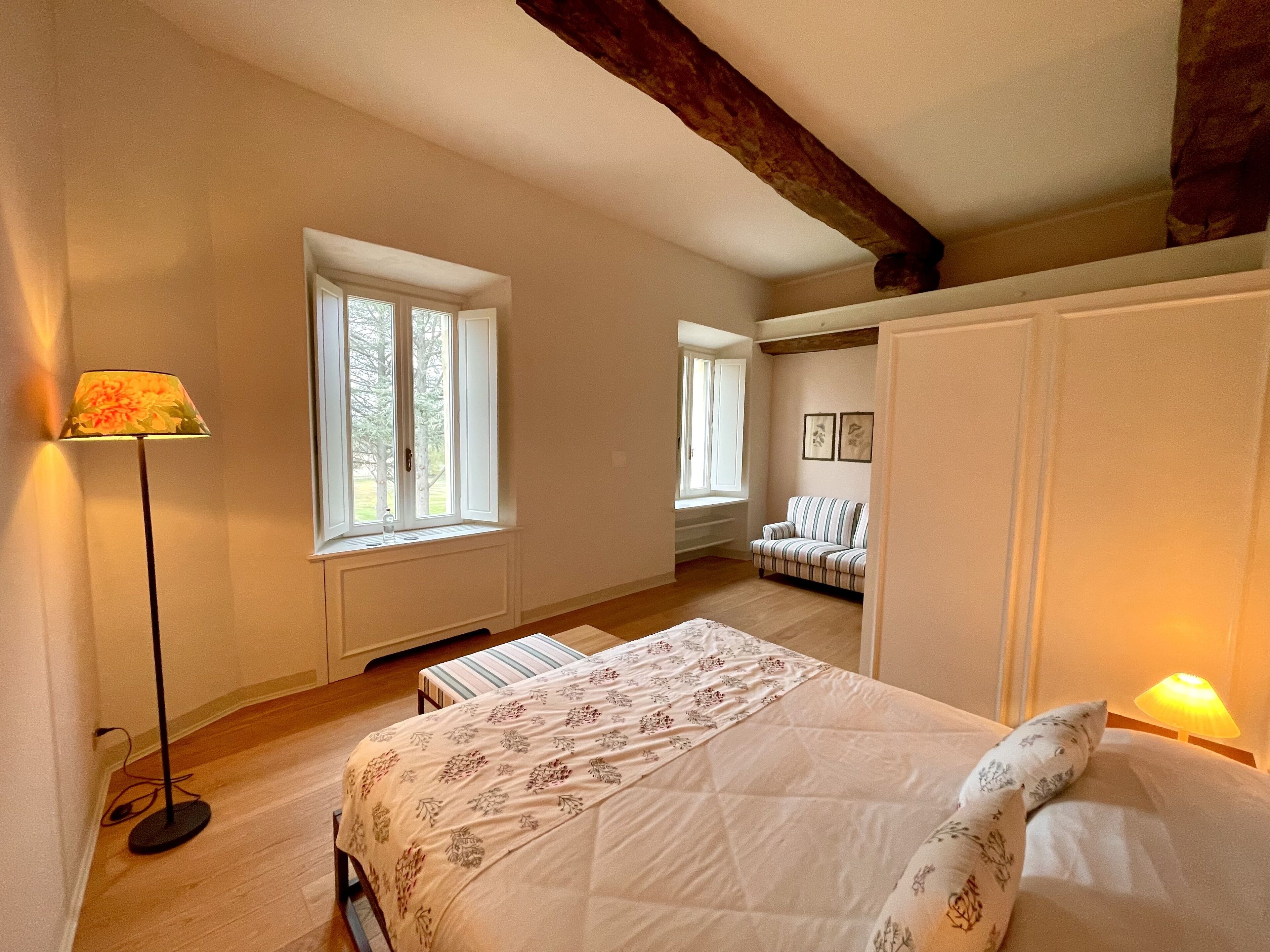 A bedroom with a bed, a standing lamp, a window, and a sofa.