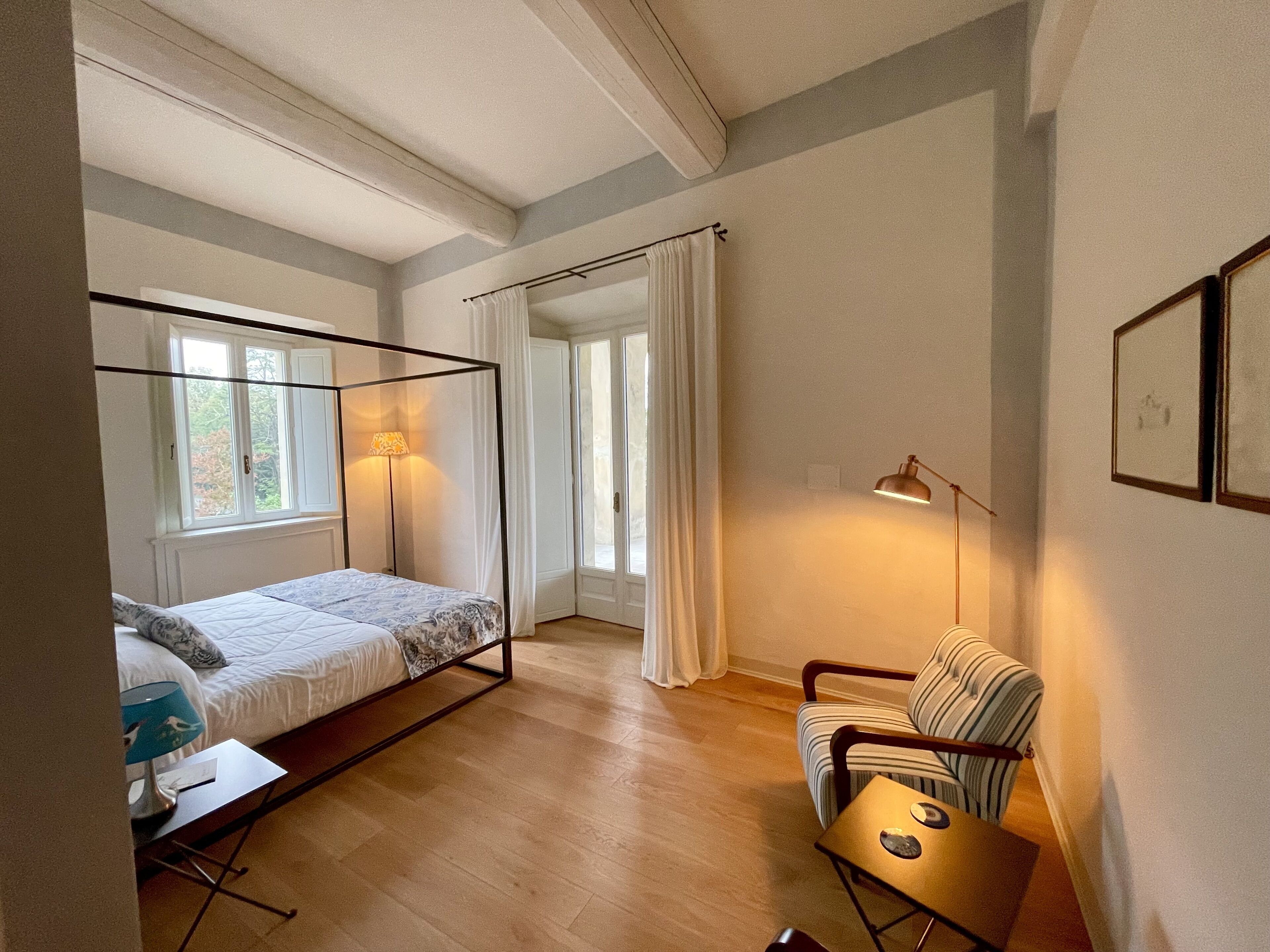 Un dormitorio con cama de dosel, una mesita de noche con lámpara, una silla y una ventana con cortinas.