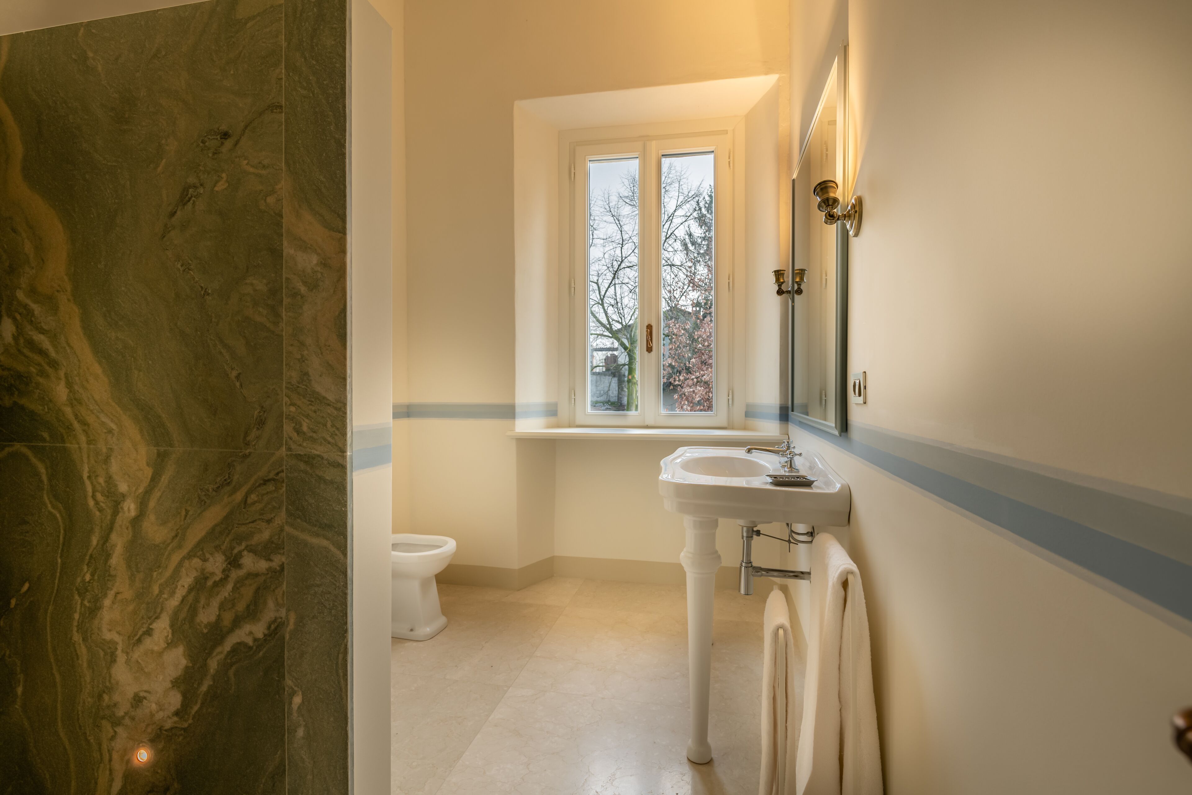 Een badkamer met een marmeren vloer, een wit wastafelmeubel, een raam met uitzicht op bomen en een toilet.