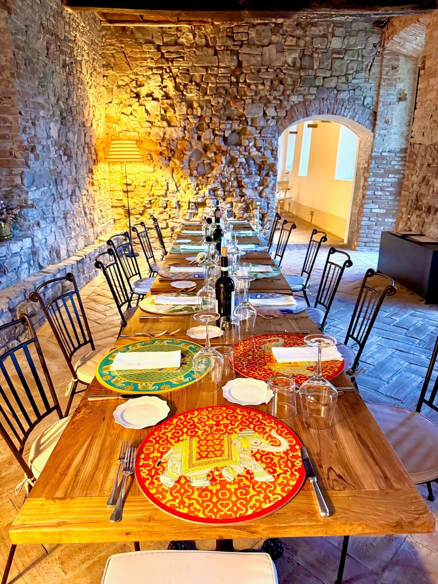 Une longue table à manger, dressée avec des assiettes, des couverts et des verres, entourée de murs en pierre et de chaises en métal.