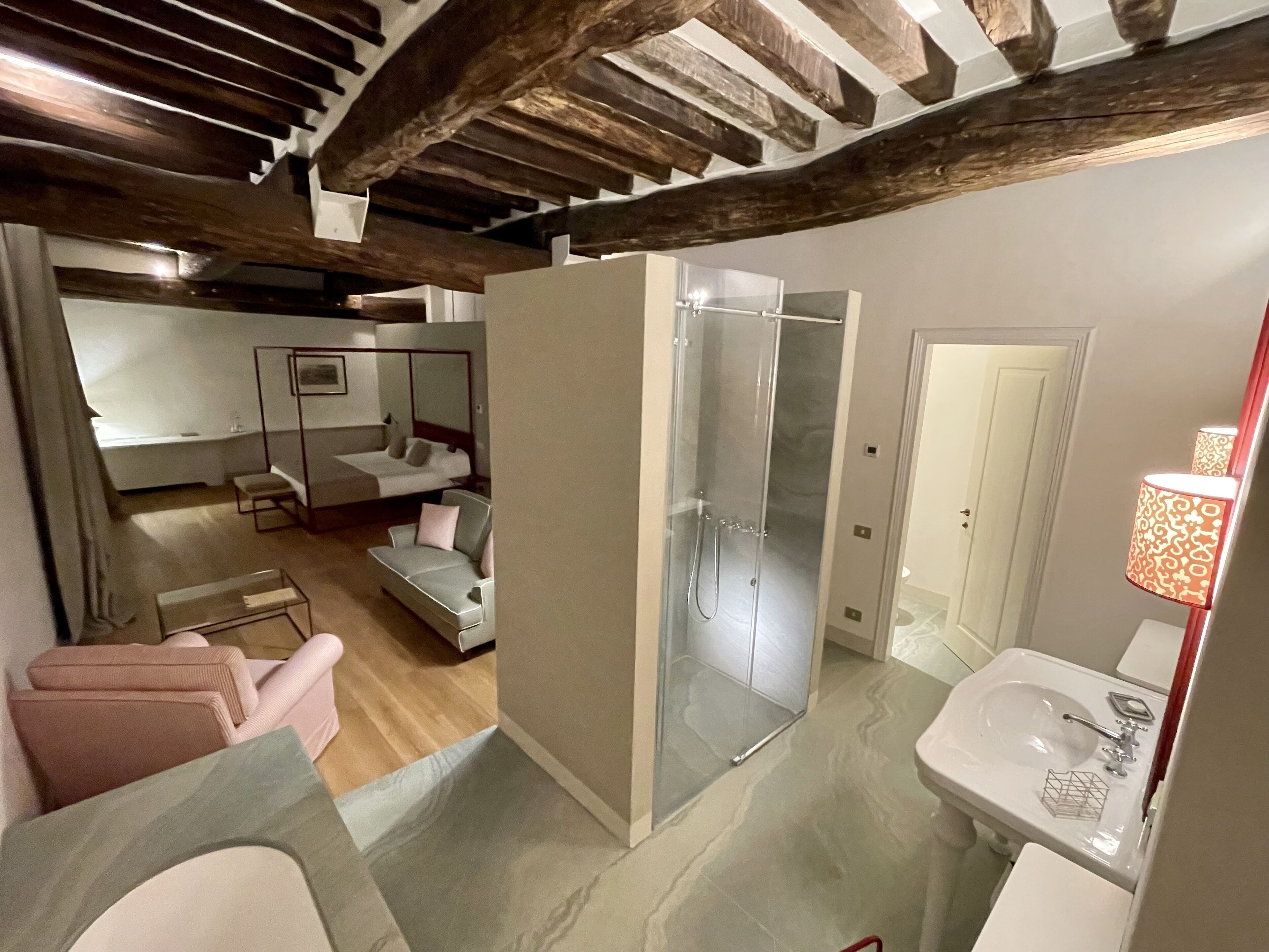 Una camera d'albergo moderna con soffitto in legno, box doccia in vetro, un letto con testiera, una zona relax con cuscini e un bagno con lavandino e specchio.