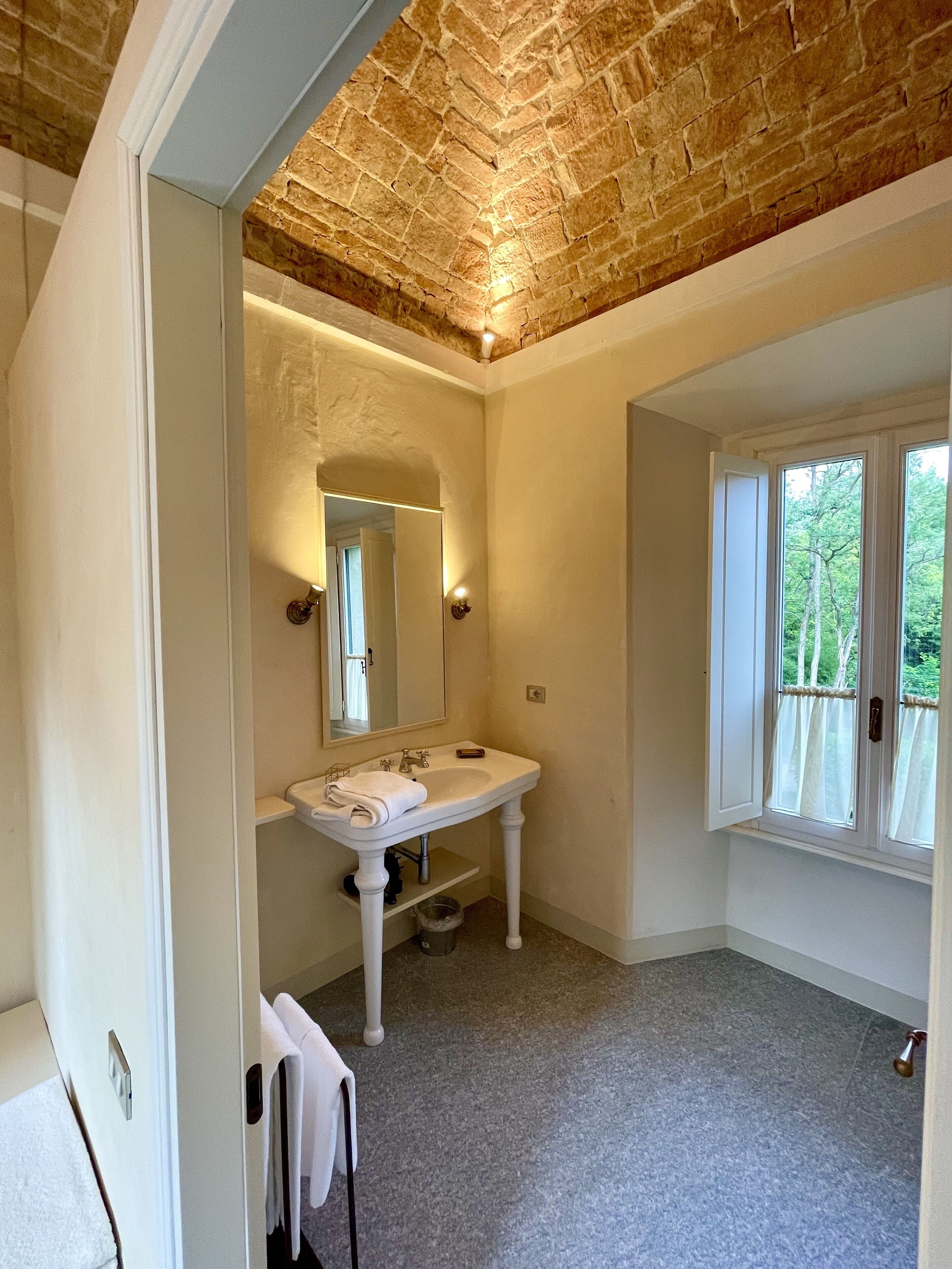 Une salle de bain avec un lavabo sur colonne blanc, un miroir au-dessus et une fenêtre avec des rideaux.