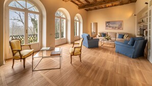 Un soggiorno spazioso con pavimento in legno, soffitto alto con travi a vista e ampie finestre ad arco.