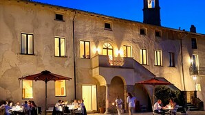 Un'area pranzo all'aperto con tavoli e ombrelloni, un edificio con una torre dell'orologio e terreno sterrato.