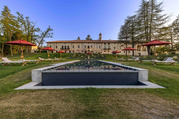 A pool with a geometric design, surrounded by lounge chairs and red umbrellas, with a large building in the background.