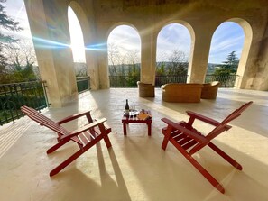 A covered outdoor seating area with red wooden chairs, a small table with a bottle and glasses, and a wicker sofa.