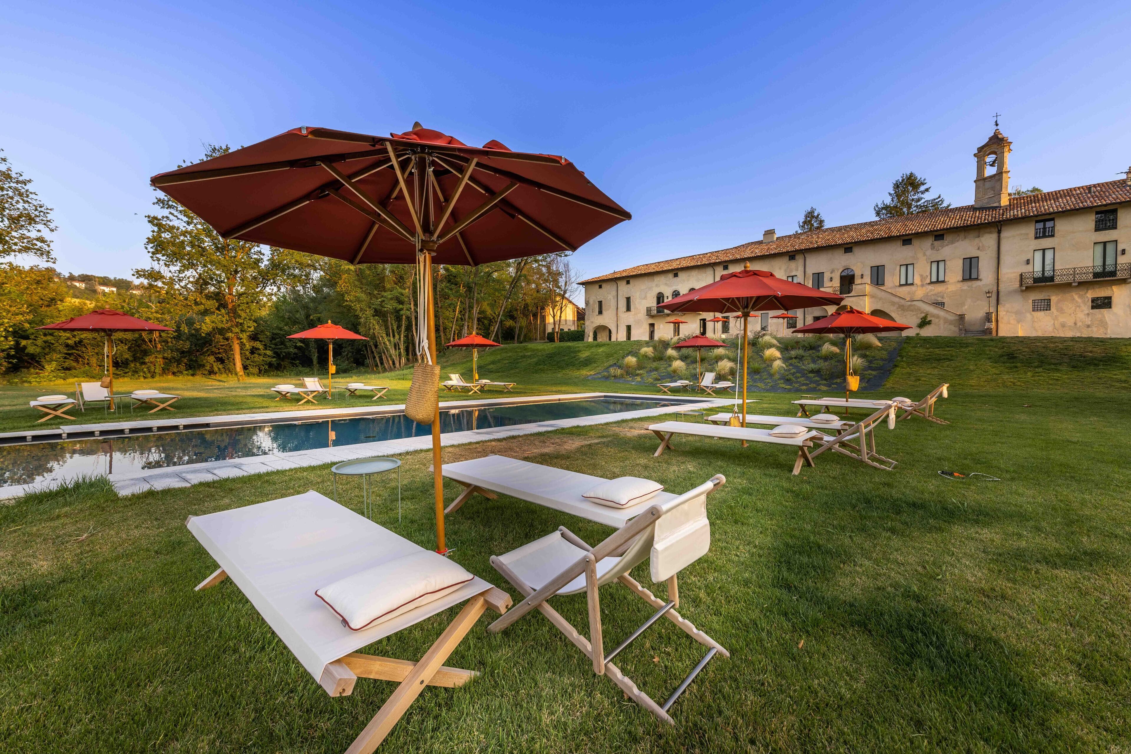 Zona junto a la piscina con sillones de descanso y sombrillas rojas, una piscina pequeña y un edificio con una torre de campanas al fondo.