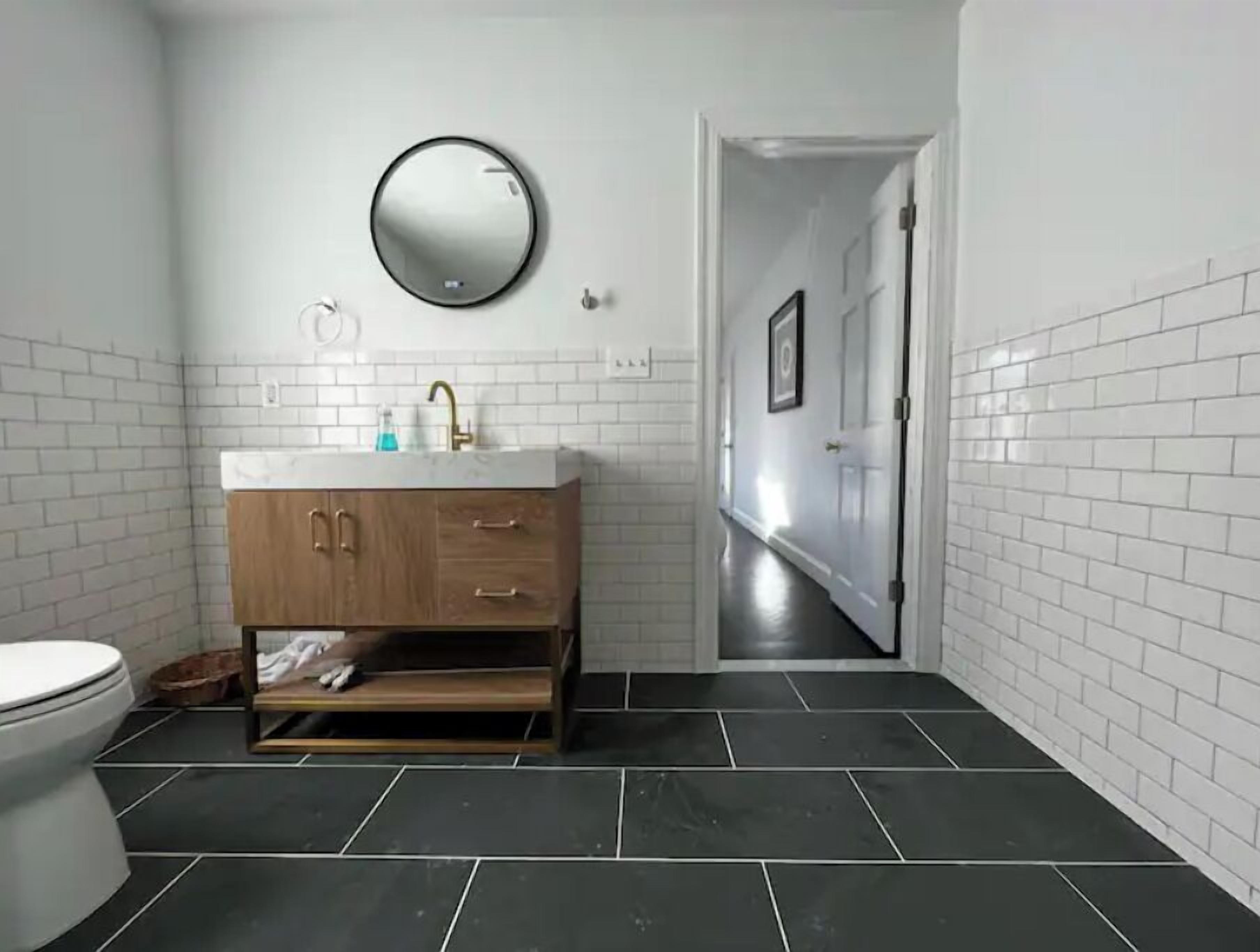 Een badkamer met een witte betegelde muur, een houten wastafelmeubel met een ronde spiegel, en een zwarte betegelde vloer.
