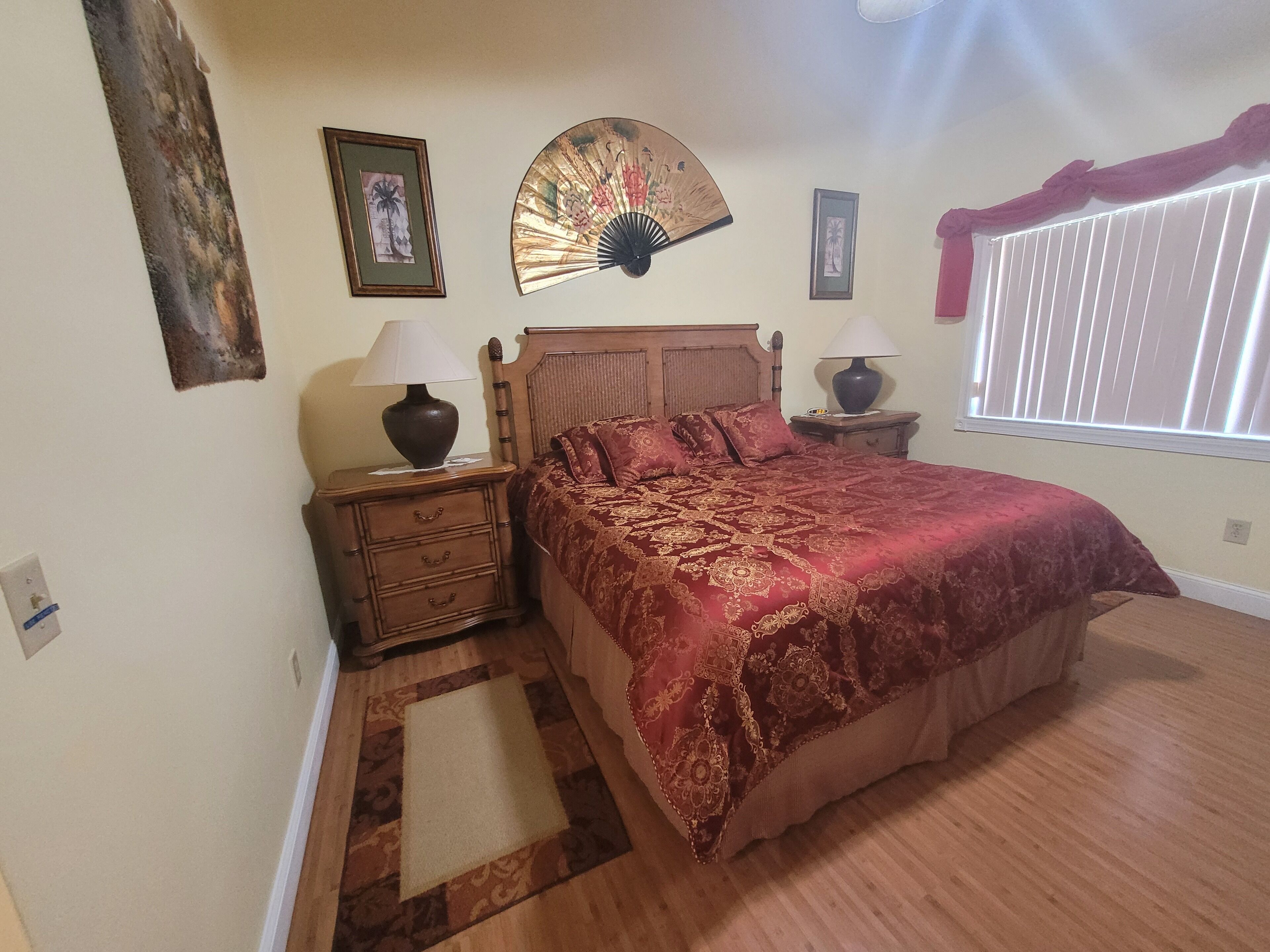 A bedroom with a wooden bed, two bedside tables with lamps, a decorative fan, and a window with blinds.