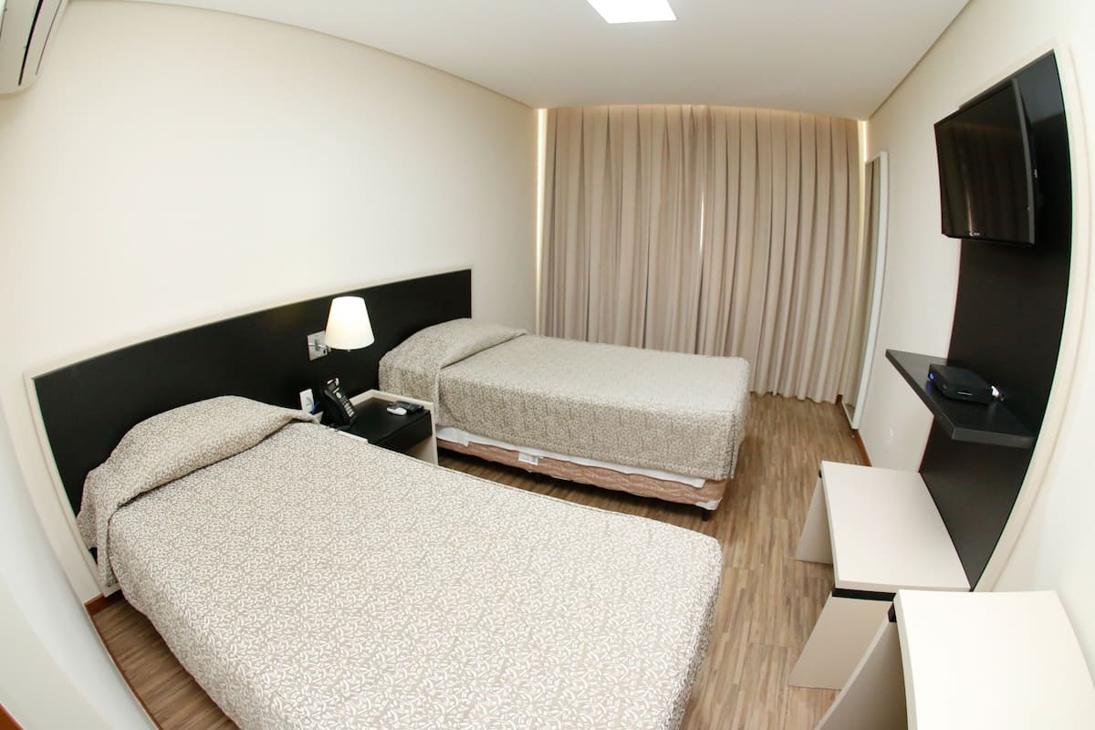 A hotel room with two beds, a desk, a TV, and a window with curtains.