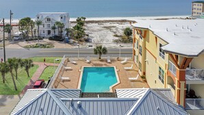 Um hotel com piscina no terraço, vista para a praia e área de estacionamento com um carro vermelho.