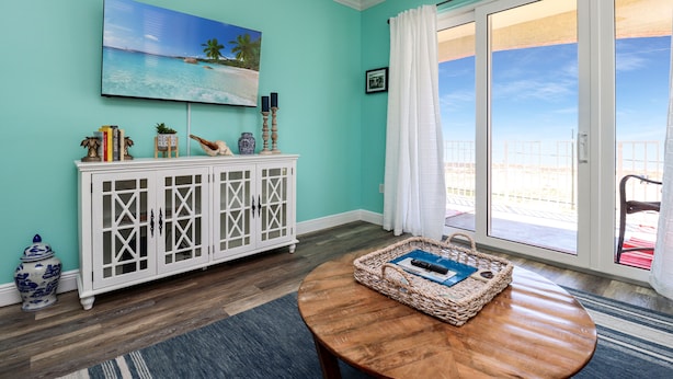 Quarto com paredes verde-água, um armário branco, uma mesa de madeira redonda e uma grande janela com vista para a praia.