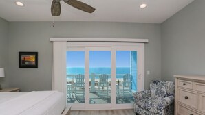 A bedroom with a bed, a chair, a dresser, and a sliding glass door leading to a balcony with chairs and a view of the ocean.