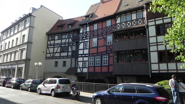 Un bâtiment traditionnel à colombages, avec plusieurs étages et des balcons, des voitures garées le long de la rue et un trottoir pour les piétons.