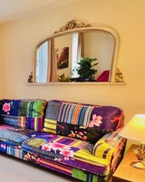 Un sofá colorido con diseño de patchwork, un espejo grande y ornamentado arriba, y una lámpara sobre una mesita de noche.