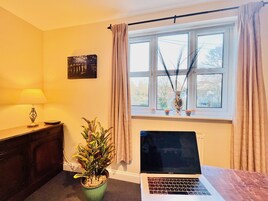 Una habitación con un ordenador portátil sobre un escritorio, una planta en maceta, una lámpara y una ventana con cortinas.