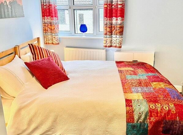 A neatly made bed with a white comforter and red pillows, a colorful quilt, and a window with patterned curtains. - Dapps Hill Haven (Bristol)